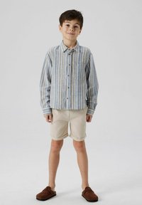 Chemise à rayures bleu, beige et blanc, avec une poche ; shorts couleur crème ; chaussures slip-on brunes ; textures de tissus légers.