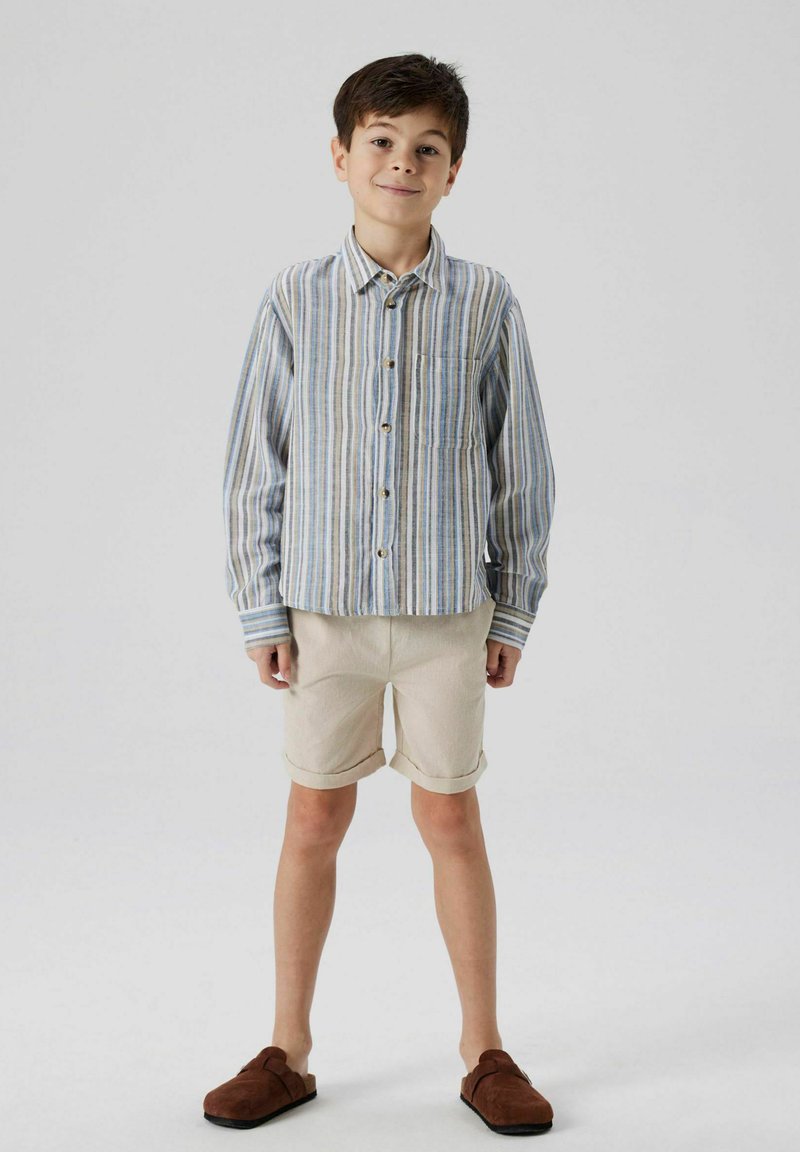 Chemise à rayures bleu, beige et blanc, avec une poche ; shorts couleur crème ; chaussures slip-on brunes ; textures de tissus légers.