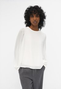 Blusa blanca con mangas largas transparentes y abullonadas, escote redondo y textura suave, combinada con pantalones grises ajustados que presentan un sutil estampado de rayas.