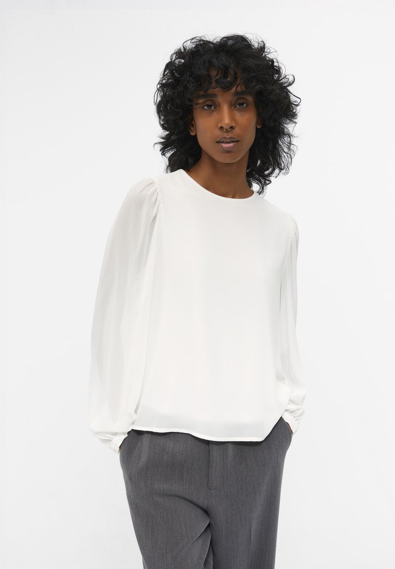 Blusa blanca con mangas largas transparentes y abullonadas, escote redondo y textura suave, combinada con pantalones grises ajustados que presentan un sutil estampado de rayas.