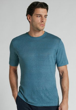 Hombre con cabello castaño que lleva una camiseta azul de manga corta y cuello redondo, de pie con las manos en los bolsillos, mirando de lado sobre un fondo liso.