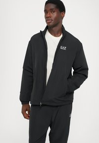 Black, lightweight jacket with a zipper and elastic cuffs. Features a small white logo on the left chest and a relaxed fit.