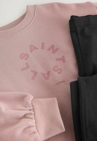 Roza sweatshirt z okroglim logotipom "SAINT". Ima rebrast izrez in nabran detajl rokavov. Črne hlače so zložene ob strani.