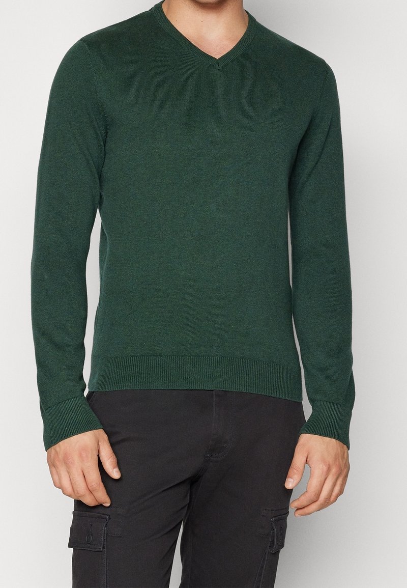 Pull vert à col en V avec poignets et ourlet côtelés. Texture douce et coupe ajustée. Porté avec un pantalon cargo noir.