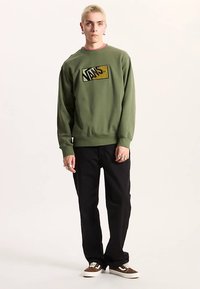 Grön sweatshirt med en rektangulär Vans-logotyp i svart och guld. Bärs med svarta byxor och bruna sneakers, med en avslappnad passform.