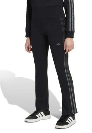 Image recadrée d’un modèle portant un pantalon de survêtement noir Adidas évasé, à bandes latérales, avec des baskets noires assorties à bandes blanches.