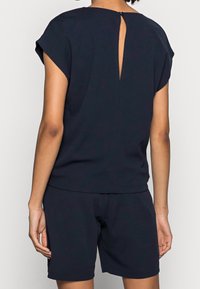 Kobieta w czarnej bluzce z krótkim rękawem z wycięciem i guzikiem z tyłu oraz pasujących czarnych szortach, ukazana od tyłu na jednolitym tle.