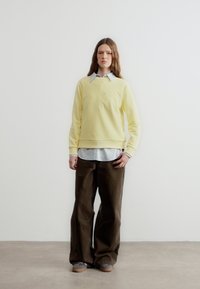 Άτομο που στέκεται μπροστά σε λευκό τοίχο φορόντας κίτρινο φούτερ, ριγωτό πουκάμισο με γιακά, φαρδιές καφέ παντελόνες και γκρι παπούτσια, με ουδέτερη έκφραση.