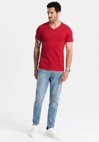 Rød kortærmet V-hals t-shirt, parret med lyseblå jeans og hvide sneakers. Modellen står frontalt mod en ensfarvet baggrund.