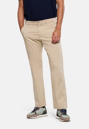 Hombre con pantalones beige de pierna recta y zapatillas verdes, grises y marrones con cordones azul marino, de pie con una mano en el bolsillo.