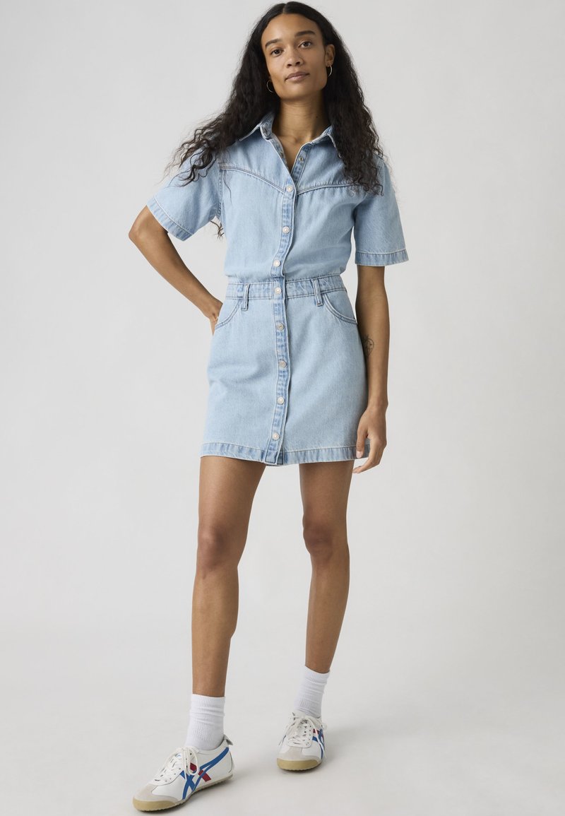 Femme aux longs cheveux bouclés portant une robe chemise en jean bleu clair, des chaussettes blanches et des baskets blanches avec des rayures bleues et rouges.