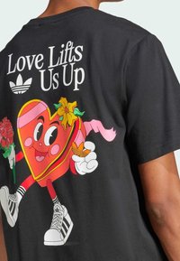 T-shirt en coton noir arborant un personnage de cœur coloré tenant des fleurs. Le texte au-dessus indique "L'amour nous élève" avec le logo Adidas.