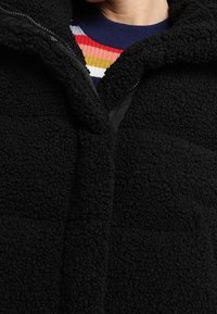 Sort fleecejakke med en tekstureret overflade, der har en frontlynlås og en høj krave. Under den er der en flerfarvet stribet sweater.