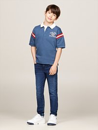 Polo blu con strisce rosse e bianche sulle maniche, logo sul davanti, abbinato a jeans scuri e sneakers bianche.