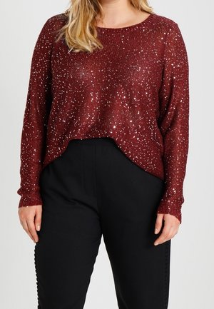 Kvinde iført en langærmet vinrød sweater med sølvglimmer og sorte bukser, stående mod en ensfarvet lys baggrund.