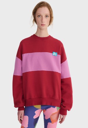 Młoda osoba z rudymi włosami, ubrana w czerwoną i różową paskowaną bluzę oraz kolorowe kwieciste leginsy, stojąca na tle jasnej, jednolitej ściany.