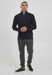 Marineblauer Strickpullover mit Reißverschlusskragen, strukturiertem Stoff, kombiniert mit grauen schmal zulaufenden Hosen und schwarzen Sneakers mit weißer Sohle.