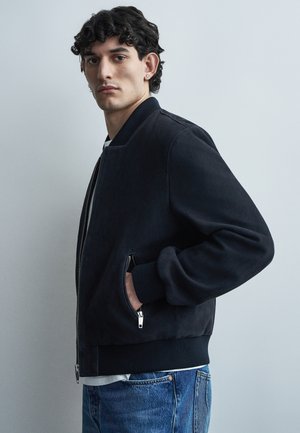 Jeune homme aux cheveux bouclés portant une veste bomber noire zippée et un jean bleu, debout avec une main dans la poche devant un fond uni.