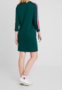 Grünes Sweatshirtkleid mit Dreiviertelärmeln, das rote, weiße und marineblaue Streifen an den Schultern aufweist. Kombiniert mit weißen High-Top-Sneakern.