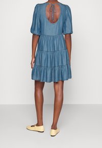 Vestido de denim con mangas cortas abullonadas, detalle de lazo en la espalda abierta y dobladillo con volantes en capas. El color es azul claro. Combinado con zapatos planos amarillos.