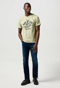 Jasno oliwkowa koszulka z krótkim rękawem z czarnym graficznym tekstem i logo, połączona z ciemnoniebieskimi dżinsami i czarnymi butami. Wygodne dopasowanie.