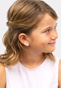 Silver heart-shaped earring with a textured finish, set against light brown waves of hair. The background is a plain, light color.