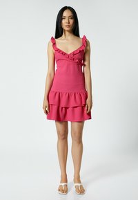 Robe rose ajustée avec des bretelles volantes, design enroulé et jupe à étages. Fabriquée en tissu texturé avec un léger éclat.