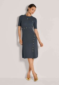 Frau in einem knielangen dunkelgrauen Kleid mit goldenen Knopfakzenten und goldenen High Heels, die vor einem schlichten hellen Hintergrund posiert.