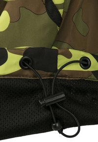 Kamouflage-tyg med gröna och bruna mönster samt en svart nätsektion nedanför. Inkluderar en svart dragsnodd med en justerbar klämma i botten.