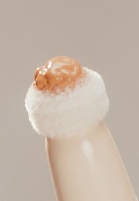 Aplicador de maquillaje con una punta redonda, suave y blanca y producto marrón encima. El mango es beige y de textura suave.