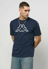 Mann med kort hår iført en marineblå t-skjorte med en hvit silhuettlogo av to personer som sitter rygg mot rygg, og lyseblå jeans.