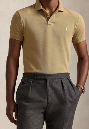Homme portant un polo beige avec un logo blanc et un pantalon plissé gris foncé taille haute, montre au poignet gauche, main dans la poche.