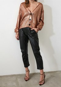 Cárdigan de punto en rosa dorado brillante con un escote en V profundo y grandes botones negros, combinado con pantalones ajustados de cuero sintético negro y tacones atados.