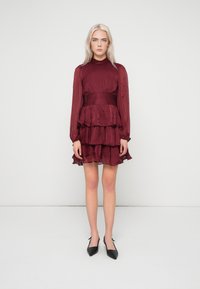 Γυναίκα με μακριά ξανθά μαλλιά, φορώντας ένα μπορντό φόρεμα με ψηλό λαιμό και επίπεδες στρώσεις, με μακριά διάφανα μανίκια και μαύρες γόβες με μυτερή μύτη, που στέκεται σε λευκό φόντο.