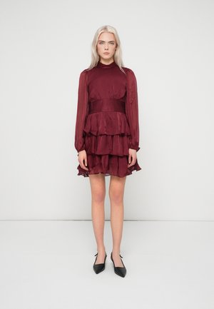 Kvinna med långt blont hår som bär en bourgogne höghalserad klänning med flera lager, långa genomskinliga ärmar och svarta spetsiga klackar, stående på vit bakgrund.