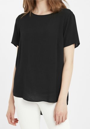 Femme portant un chemisier noir ample à manches courtes associé à un pantalon blanc moulant sur un fond uni.