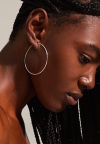 Boucle d'oreille en argent, grande taille, accrochée à une oreille à la peau foncée avec des cheveux tressés. La texture de la peau est lisse, avec un éclairage doux mettant en valeur les détails.