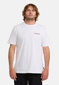 Mann mit hellbraunen Haaren, der ein weißes Billabong-T-Shirt mit kurzen Ärmeln und schwarze Hosen trägt, steht vor einem schlichten weißen Hintergrund.