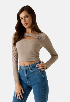 Giovane donna con lunghi capelli castani che indossa un top corto beige con dettagli in strass e jeans blu a vita alta, in posa con una mano sul fianco.