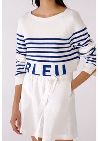 Pull en tricot blanc avec des rayures horizontales bleues, arborant le mot "BLEU" à l'ourlet. Associé à un short blanc, mettant en avant une coupe décontractée.