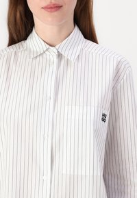 White button-up shirt with vertical black stripes, classic collar, and left chest pocket featuring "HUGO" logo in black stitching.