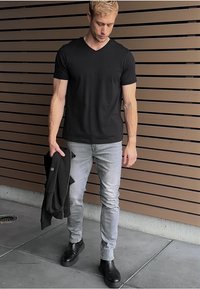 Zwarte v-hals t-shirt gecombineerd met strakke lichtgrijze jeans, zwarte enkellaarsjes, en een donker jack dat over één arm is gedrapeerd. Achtergrond van houten panelen.