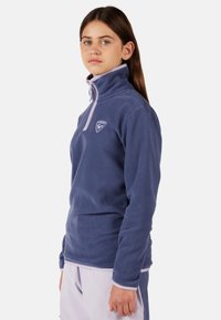 Modrý fleecový pulovr s vysokým krkem, předním zipem do poloviny a kontrastním levandulovým lemem. Má logo na hrudi a dlouhé rukávy.