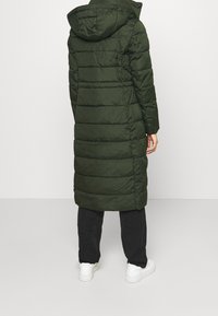 Długi, pikowany płaszcz z kapturem w ciemnozielonym kolorze. Charakteryzuje się pionowym przeszyciem i taliowaniem. Połączony z ciemnymi spodniami i białymi trampkami.