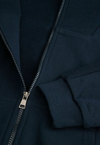 Ciemnoniebieska kurtka na zamek z wewnętrznym polarem, z ściśniętymi mankietami i metalowym zamkiem. Gładki materiał z teksturowaną wstawką na ramionach.