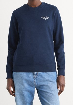Person trägt einen dunkelblauen Sweatshirt mit langen Ärmeln und dem Schriftzug "Tommy Jeans New York City" auf der Brust sowie hellblaue Jeans.