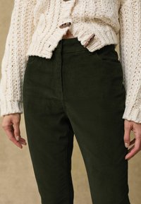 Pantaloni in corduroy verde scuro con una morbida trama, vita alta e sottili linee verticali, abbinati a un cardigan bianco lavorato a maglia spesso.