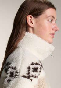 Kurtka z polaru z wysokim kołnierzem, w kolorze kremowym, z brązowym wzorem; materiał o fakturze, miękki; zamek błyskawiczny przy szyi.