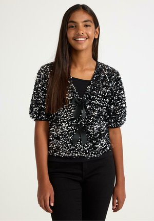 Černý kardigan s flitry a krátkými nadýchanými rukávy, zdobený dvěma černými stuhovými mašlemi vpředu. Kombinován s černým topem a kalhotami.