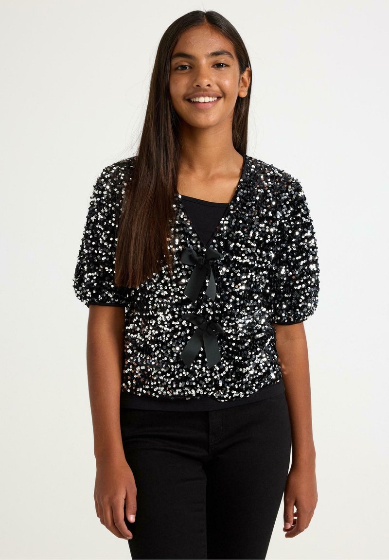 Černý kardigan s flitry a krátkými nadýchanými rukávy, zdobený dvěma černými stuhovými mašlemi vpředu. Kombinován s černým topem a kalhotami.